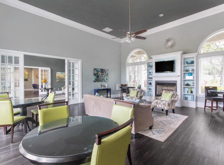 Clubhouse Seating Area with Large Windows on either side of Fireplace with TV and Shelves Surrounding Fireplace Multi Colored Accent Chairs Grey Couch and Dining Room Tables with Green Chairs and Clubhouse Foyer in the Background